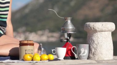 Tanınmayan bir kadın Karadağ 'da piknikte yerde oturuyor ve Donji Stoliv körfezindeki bir körfezde kamp gaz ocağında kahve yapıyor. Kadın, arka planda bulanık dağ manzaralı açık havada kahve yapıyor.