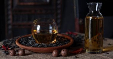 Yuvarlak cam kase, fındık ve kurutulmuş kırmızı biberlerle çevrili siyah tohumlarla dolu ahşap bir tabağa yerleştirilmiş sıcak yağ içerir. Toprak ve kırsal bir atmosfer yaratır..