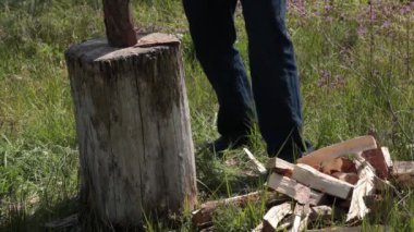 Adam yakacak odun kesiyor, yakındaki yeşil çimlerin üzerinde yakacak odun yığını var. Odun balta darbesiyle kırılır ve düşer. Oduncu kır şöminesi için odun kesiyor.