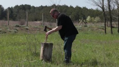 Siyah tişörtlü ve güneş gözlüklü orta yaşlı vasıfsız bir adam kırsal alanda, bahçenin dışında küçük bir baltayla yakacak odun kesmeye çalışıyor. Yakacak odunları uygunsuz bir şekilde kesiyor.