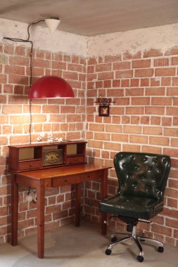 Dark vintage chair on wheels against brick wall next to wooden table on which there is shelf with old clock over which large red lamp hangs. Interior of retro room. Vintage style interior clipart