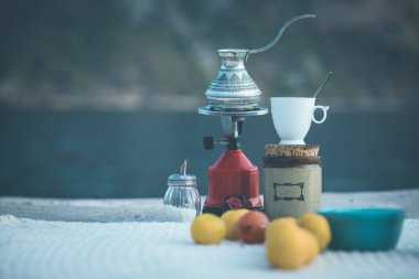 A vibrant coffee pot sits on a small stove, surrounded by fresh lemons, a rustic jar, and a white mug with a spoon. clipart