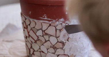 Kırık fayanslı mozaik saksı tasarlama süreci. İnsan, macunu spatulayla süsleyerek saksıyı mozaikle süslüyor. Bahçe süslemeleri için DIY el işi projesi