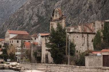Eski taş yapılar, mimari tarzların bir karışımını sergileyerek rıhtımı kaplıyor. Karadağ 'ın sakin kıyı cazibesini vurgulayarak, arka planda kayalık tepeler belirgin şekilde yükseliyor 
