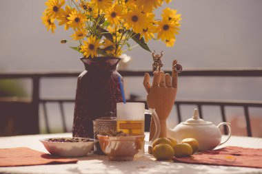 Canlı bir masa düzeninde sarı çiçeklerle dolu bir vazo, çeşitli fındık kaseleri, bir çaydanlık ve ayırt edici ahşap bir el heykeli sergilenir. Sıcak güneş ışığı davetkar atmosferi güçlendirir..