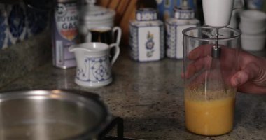 Plastik bardağı yumurta ve şekerin sarı karışımından tut. Yumurtaları şekerle blender ile karıştırın. Köpük ortaya çıktı. El ele tutuşan konteynır. Mutfakta mutfak gereçleri var. Yemek pişirme aşaması