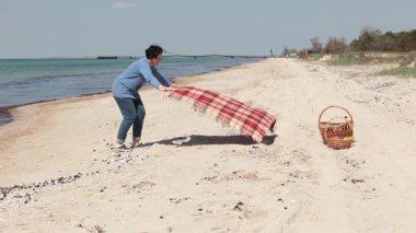 Kadın plajda piknik yapmak için kumsala ekose battaniye serer. Dalgalar kıyıları, deniz kıyısını, deniz manzarasını yıkar. Açık hava eğlencesi, plajda piknik..