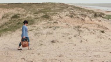 Kadın piknik yapmak için kumlu sahilde sepetler dolusu yiyecekle yürüyor, kamera kadını takip ediyor. Dalgalar kıyıya vurur, deniz kıyısı doğası, deniz manzarası. Açık hava eğlencesi, deniz kıyısında yürümek, sahilde piknik..