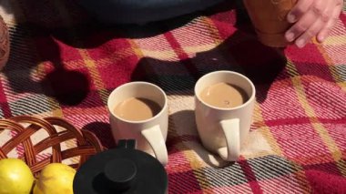 Canlı bir piknik battaniyesinin etrafında toplanan arkadaşlar, açık havada taze meyvelerin tadını çıkarırken sıcak içeceklerin tadını çıkarıyorlar. Güneş neşe dolu atmosfere katar ve kalıcı anılar yaratır..