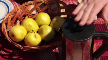 Fransız presinden kupalara kahve dök, kil testisinden süt ekle ve şeker koy. Piknikte kahve yapma süreci. Kırsal tarzda piknik. Kareli battaniyede örülü sepetlerle hayat.