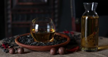 Cam ve ahşap bir tabak ayçiçeği tohumu ve bir şişe altın tohum yağı. Pişirme yağı hala hayat dolu. Ön planda kesilmiş fındıklı Macadamia fıstığı. Ay çekirdeği arka plandaki masaya düşer.