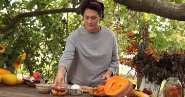Bir kadın Şükran Günü için hazırlanıyor. Balkabağı ve böğürtlen gibi taze malzemeleri yeşil bir masa üzerinde dikkatlice hazırlıyor. Açık havada canlı bir atmosfer yaratıyor..
