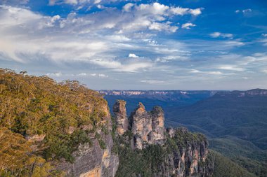 Avustralya 'nın Yeni Güney Galler bölgesinin pitoresk mavi dağlarında bereketli yeşillikler ve mavi gökyüzü ile çevrili ikonik üç kız kardeş kaya oluşumunun nefes kesici bir görüntüsü, akşam güneşinin sıcak renklerinde güneşleniyorlar..