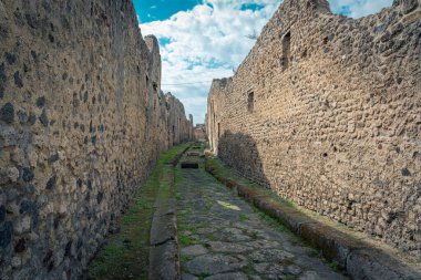 İtalya 'nın Pompeii kentindeki yıpranmış taş duvarlar ve kalıntılarla çevrili antik taş patika. Site, antik mimariye bir bakış sunan canlı mavi gökyüzü altında dikkate değer tarihi bir korunmayı ortaya koyuyor..