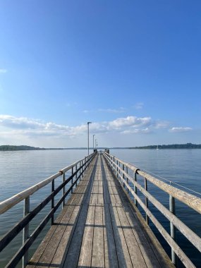 Bavyera 'daki Berrak Göl Altındaki Sessiz Mavi Göl Chiemsee' ye uzanan Uzun Ahşap İskele. Berrak bir gökyüzünün altında sakin bir mavi göle uzanan, huzuru çağrıştıran ve kaçmayı başaran ahşap bir iskelenin sakin bir görüntüsü.