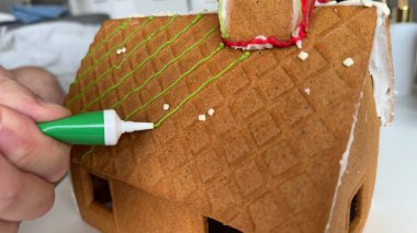 Elle dekore edilen zencefilli ev çatısı, Yeşil Buzlu Şekerleme ile Şenlik Dokunuşu. Birisinin zencefilli kek evinin çatısına taze yeşil krema sürmesine yakın çekim. Kutlama dekorasyonları arasında krema da var