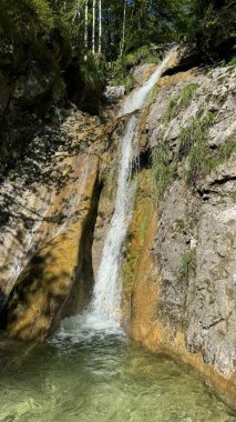 Scenic Waterfall Cascading Over Rocky Cliff Surrounded by Lush Green Trees in Berchtesgaden, Germany. A serene natural waterfall flows gracefully over a rocky cliff into a clear pool and than into a clipart