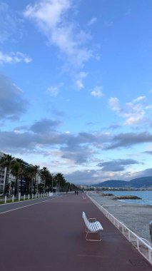 Nice 'teki Fransız gezinti güvertesinde huzurlu bir yürüyüş. Fransa 'nın Nice kentindeki Gezinti Güvertesi' nde sakin bir akşam yürüyüşü. Beyaz banklar, yol boyunca palmiye ağaçları, manzaralı bir manzara.