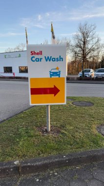 22.03.2025 Almanya: Kabuk Benzin İstasyonu Araba Yıkama İşareti ve Yön Oku. Bir Shell araba yıkama tabelası çimlerin üzerinde duruyor, parlak ok işaretli araçlara yön veriyor..