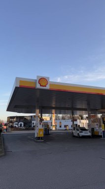 22.03.2025 Almanya: Günbatımında Logolu Modern Benzin İstasyonu Kabuğu. Açık bir gökyüzü altında logosu olan çağdaş bir Shell benzin istasyonu, beyaz bir park etmiş araba ve çeşitli yakıt ikmali sergiliyor.