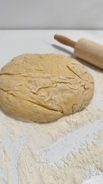 Taze hazırlanmış hamur un kaplı bir yüzeyde duruyor. Yanında merdane var. Çeşitli pişirme faaliyetleri için idealdir.