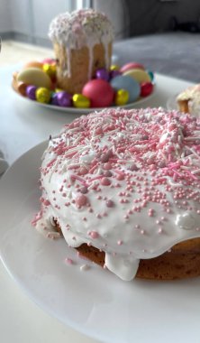 Pembe süslemeli ve beyaz kremalı pasta canlı paskalya yumurtalarıyla çevrili bir tabakta oturur. Bahar kutlamaları ve toplantılar için mükemmel neşeli ve şen bir atmosfer yaratır.