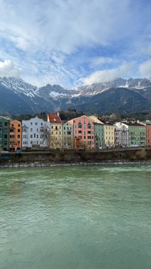 Innsbruck, Avusturya - 28 Mart 2025: Büyüleyici pastel renkli binalar, bir nehir kenarı Inn 'i karla kaplı Alp dağları ve açık mavi gökyüzü manzarası sunuyor