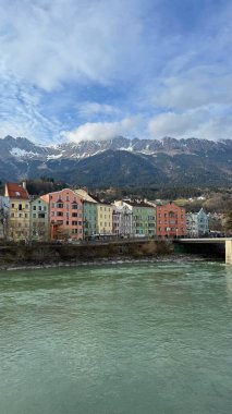 Innsbruck, Avusturya - 28 Mart 2025: Riverside evleri canlı renklerle boyandı, karla kaplı dağların manzaralı zeminleri ve önplanda sakin bir nehir hanı vardı.
