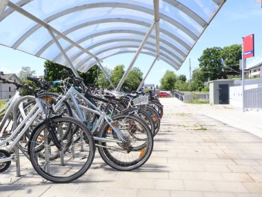 31.05.2025 Almanya: Mavi Gök ile güneşli bir günde tren istasyonunda bisikletli park alanı kaplandı. Bisikletler modern tasarlanmış bir gölgeliğin altında iyi organize edilmiş bir bisiklet park alanında park halindedir.