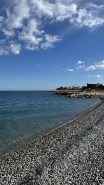 Nice, Fransa 'da kıyı manzarası, kıyı şeridi boyunca çakıl taşları ve parlak bir gökyüzünün altında berrak mavi su, rahatlama ve tatil için ideal doğal güzellik ve sükunetin özünü yakalıyor.