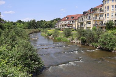 Nehir kenarındaki evlerden akan dere kırsal alanda güneşleniyor. Sakin bir Mangfall deresi, güneşli bir gökyüzünün altındaki konut binalarının yanındaki yoğun yeşilliklerden akıyor. Huzurlu bir havayı çağrıştırıyor.