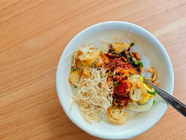 A bowl of traditional rice porridge topped with shredded chicken, fried shallots, peanuts, crackers, soy sauce, and garnished with fresh herbs, served on a wooden table. clipart