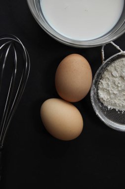 Taze yumurta, tabakta süt, süzgeçte un ve arka planda yayılmış çırpı, farklı açılarda, koyu bir arka planda yatıyor.
