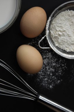 Taze yumurta, tabakta süt, süzgeçte un ve arka planda yayılmış çırpı, farklı açılarda, koyu bir arka planda yatıyor.