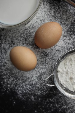 Taze yumurta, tabakta süt, süzgeçte un ve arka planda yayılmış çırpı, farklı açılarda, koyu bir arka planda yatıyor.