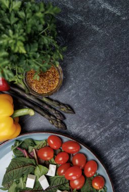 Koyu arkaplanda makarna pişirmek için malzemeler. Üstünde taze sebze salatası ve beyaz peynir tabağı olan koyu taştan bir masanın dikey fotoğrafı. Yanında dolmalık biber, kuşkonmaz, hardal tohumu, üst manzara.