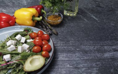 Beyaz bir masada taze sebzeler, sağlıklı vejetaryen yiyecekler. Üzerinde taze sebzeler ve beyaz peynirden salata bulunan karanlık bir taş masanın yatay fotoğrafı. Koyu bir yüzeyde salata, beyaz peynir küpleri ve yeşillikler.