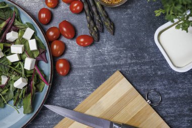 Arugula salatası, peynir, domates, soğan, zeytinyağı ve baharatlar beyaz bir masada. Sağlıklı beslenme ve vejetaryen gıda kavramı. Sağlıklı beslenme kavramı. Bıçaklı tahta kesmek ve salata için taze sebzeler. Kutudaki beyaz peynir 