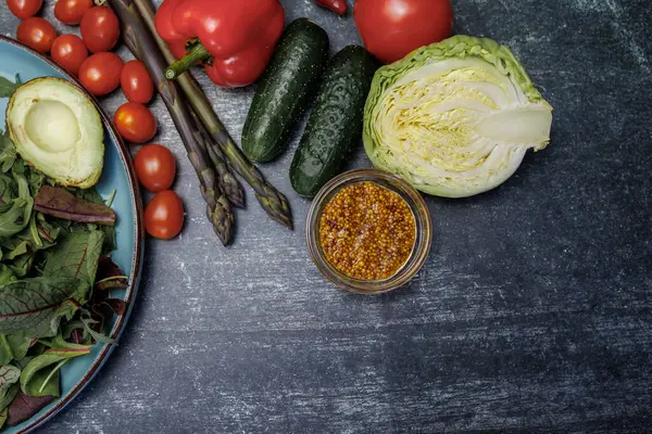 Koyu arkaplanda bir tabakta yeşil salata ve sebzeler. Koyu yüzey manzaralı bir tabakta taze sebzeler ve otlar. avokado, kiraz domatesleri, dolmalık biber, kuşkonmaz, maydanoz ve tahıllı hardal, lahana. sağlıklı beslenme endişesi