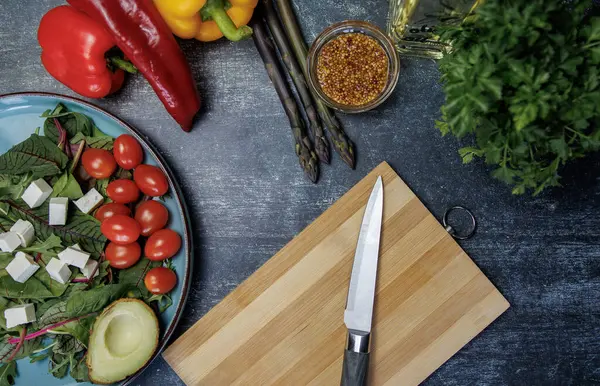 Tahta bir masada taze sebzeler. Avokado ile taze salata, kiraz domatesleri, beyaz peynir küpleri ve koyu dokulu bir yüzeyde otlar. Yakınlarda dolmalık biber, kuşkonmaz, hardal tohumu, bıçaklı ahşap tahta ve bir şişe zeytinyağı var.. 