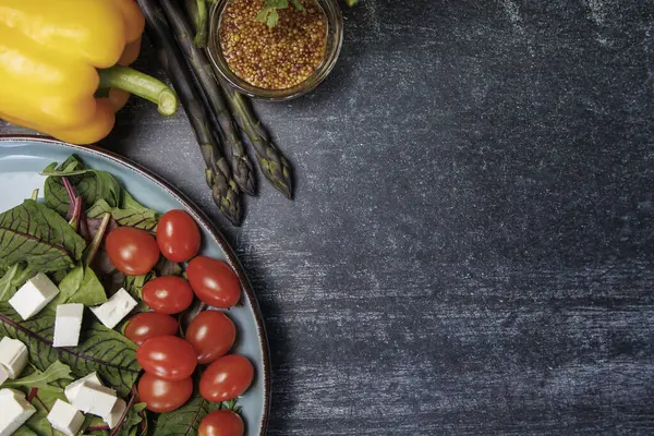 Tahta bir masada sağlıklı yiyecekler, üst manzara. Kiraz domatesli salata, beyaz peynir küpleri ve koyu bir yüzeyde yeşillikler. Çan biberi, kuşkonmaz, hardal tohumu, üst manzara, yazı için yer. konsept sağlıklı