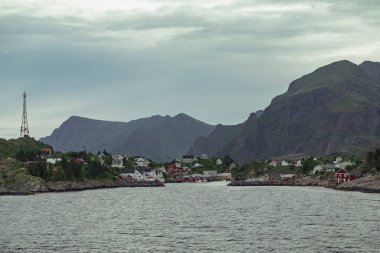 A tranquil vista of the Lofoten Islands in Norway, featuring a charming town nestled along the coast with rocky mountains rising majestically in the background under a cloudy sky. clipart