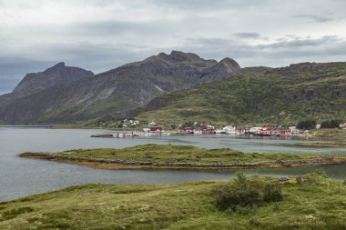 Lofoten Adaları 'nda yuva yapmış pitoresk bir köy sakin suların yanında, bulutlu bir gökyüzünün altında çarpıcı dağlarla çevrili canlı evler barındırır ve sükuneti davet eder.