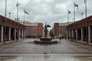 İnsanlar, Norveç, Oslo 'da bulutlarla dolu dramatik bir gökyüzünün altında modern binalar ve önemli bir heykelle çevrili canlı bir halk meydanında toplanıyorlar..