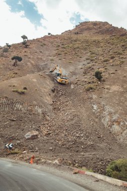 An excavator works diligently to clear debris and rubble from a steep hillside in Morocco. The rugged landscape showcases the geological features and plant life typical of this region. clipart