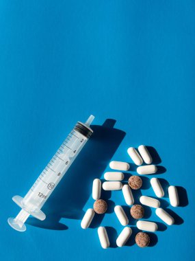 Bir şırınga, canlı mavi bir yüzey üzerinde hapların yanında duruyor. Düzenlemede beyaz kapsüller ve kahverengi yuvarlak tabletler, tıbbi tedavi ve sağlık yönetimi temalarının önerildiği.