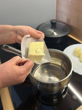 Bir insan bir parça tereyağını kağıda sarıp ocağın üstünde eritmeye hazırlanıyor. Mutfak ortamı düzenli ve temiz. Turta hazırlamak için mükemmel..