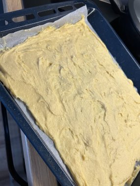 Pürüzsüz ve kremalı kek hamuru pişirmek için hazırlanmış parşömen kağıdıyla kaplı bir yemek tepsisine eşit olarak yayılır. Mutfak ayarları samimi bir pişirme oturumunu işaret ediyor..