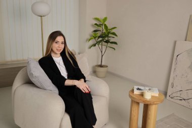 Şık siyah kıyafetli genç bir kadın, modern bir koltukta rahat bir şekilde oturuyor, yeşilliklerle dolu parlak bir iş ortamında gelişigüzel iletişim kuruyor..