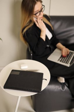 Bir kadın, dizüstü bilgisayarı üzerinde çalışırken rahat bir şekilde koltukta oturur ve bir telefon görüşmesi yapar. Yakınlarda, bir defter ve makas belirgin bir şekilde küçük bir masanın üzerine konur..